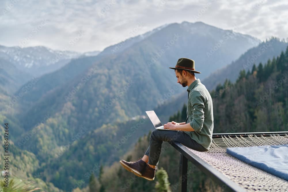 Young man freelancer traveler wearing hat anywhere working online using laptop and enjoying mountains view